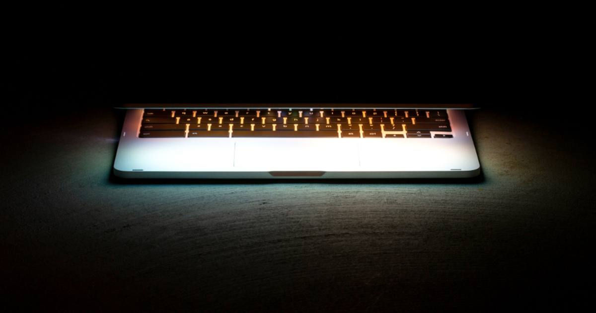 A nearly closed laptop on a dark surface, its backlit keyboard glowing through the gap